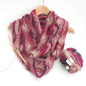 Peut inclure: Une écharpe en crochet rose, rouge et crème avec un motif de fleurs. L'écharpe est suspendue à un cintre en bois et il y a une pelote de laine à côté.