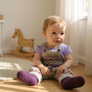 May include: A baby in a knitted romper with a lilac top and matching purple socks. The romper features a pattern in green, purple, and white. A wooden rocking horse is visible in the background. The baby is sitting on a wooden floor.