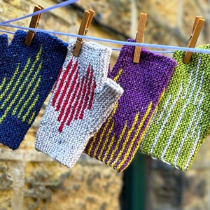 Könnte beinhalten: Vier gestrickte Fingerhandschuhe in verschiedenen Farben hängen an einer Wäscheleine. Die Handschuhe sind blau, weiß, lila und grün, jeder mit einem einzigartigen geometrischen Design in kontrastierenden Farben. Sie werden mit Holzklammern befestigt.