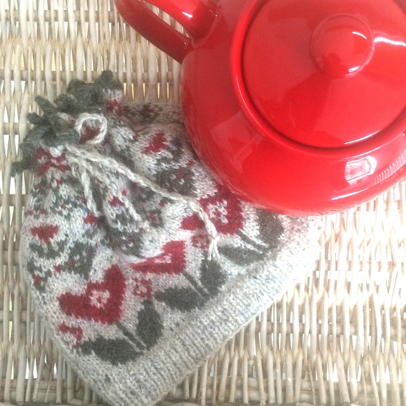 May include: A knitted hat with a white, gray, and red geometric pattern. The hat is sitting on a wicker surface next to a red teapot.