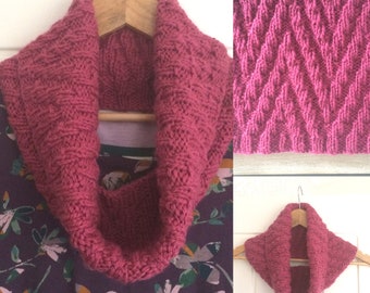 May include: A pink knitted cowl neck scarf with a textured diamond pattern. The scarf is hanging on a wooden hanger.