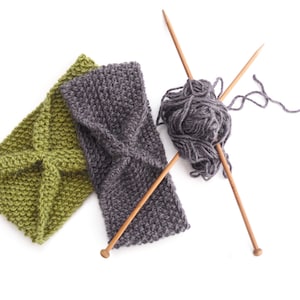 May include: Two knitted headbands, one olive green and one gray, are displayed on a white background. Knitting needles and a ball of gray yarn are also present, suggesting a handmade craft project. The headbands have a textured, woven appearance.