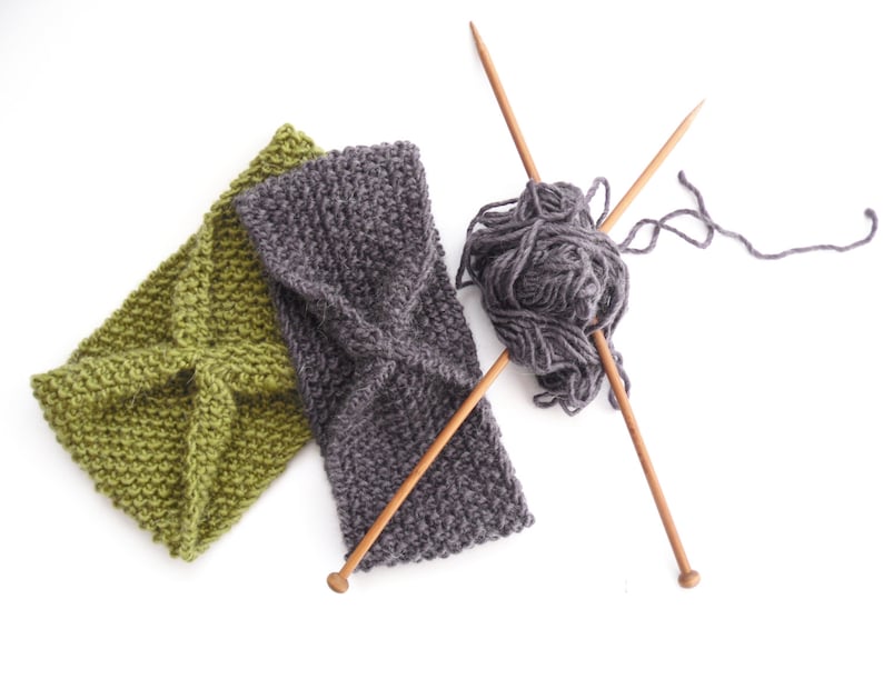 May include: Two knitted headbands, one olive green and one gray, are displayed on a white background. Knitting needles and a ball of gray yarn are also present, suggesting a handmade craft project. The headbands have a textured, woven appearance.