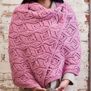 May include: A pink knitted shawl with a delicate lace pattern. The shawl is draped over a person's shoulders and is being held in place by a small metal clasp.