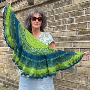 May include: A knitted shawl in shades of green and blue is displayed. The shawl features a scalloped edge and a detailed pattern. The colors are arranged in bands, creating a visually appealing design. The shawl is held up, showcasing its size and shape.