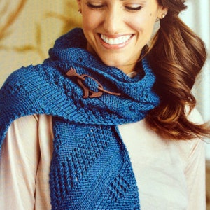 May include: A woman wearing a blue knitted scarf with a wooden kangaroo brooch. The scarf has a lacy pattern and is draped around her neck.