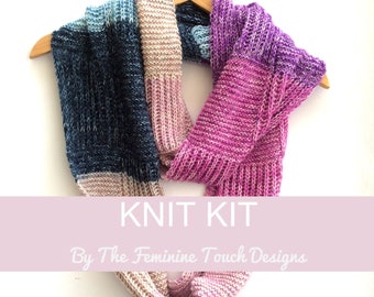 May include: Knit kit for a scarf in shades of pink, blue, and beige. The kit includes yarn and instructions. "Knit Kit By The Feminine Touch Designs" is written on a pink banner.