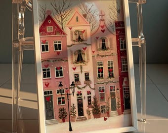 May include: A framed print featuring a whimsical illustration of several pink and red houses decorated with hearts and festive greenery. The artwork includes a street lamp and small evergreen trees, creating a charming, romantic scene.