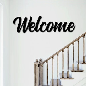 May include: Black wooden "Welcome" sign with cursive lettering mounted on a white wall above a wooden staircase.