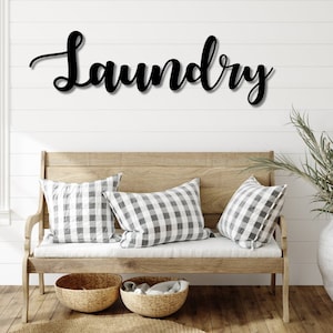 May include: A black wooden sign with the word "Laundry" in cursive script, mounted on a white wall above a wooden bench with two pillows and a woven basket.