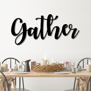 May include: Black metal wall decor with the word "Gather" in a cursive font. The decor is hanging above a dining table set with candles, a centerpiece, and glasses.