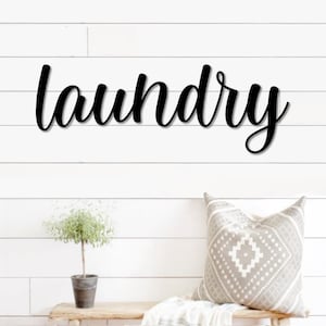 May include: Black metal sign with the word "laundry" written in a cursive font. The sign is mounted on a white wood wall.