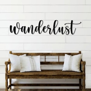 May include: Black metal wall decor with the word "wanderlust" in a cursive font. The decor is hanging on a white shiplap wall above a wooden bench with two pillows.