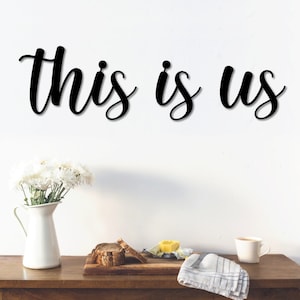 May include: Black wooden sign with the text "This is Us" in a cursive font. The sign is hanging on a white wall above a wooden table with a white pitcher, flowers, and a cup.