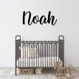 May include: Black metal name sign with the name "Noah" in a cursive font. The sign is mounted on a white wall above a gray metal crib with a white mattress and a beige pillow.