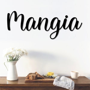 May include: Black wooden sign with the word "Mangia" written in cursive script. The sign is hanging on a white wall above a wooden table with a white pitcher, flowers, and a cup.