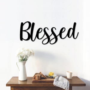 May include: Black metal wall decor with the word "Blessed" in a cursive font. The decor is hanging on a white wall above a wooden table with a vase of white flowers, a pitcher, and a cup.