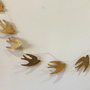 May include: A garland of decorative swallows in various shades of gold and brown. The birds are strung together on a thin wire, creating a whimsical and artistic display. The swallows are arranged in a curved line against a white background.