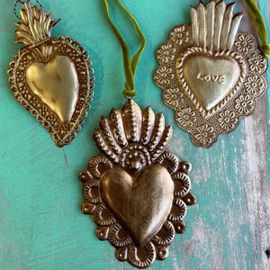 May include: Three antique gold-colored heart ornaments with intricate detailing and green velvet ribbon hangers. One ornament has the word "LOVE" inscribed. The ornaments are displayed on a distressed turquoise surface.
