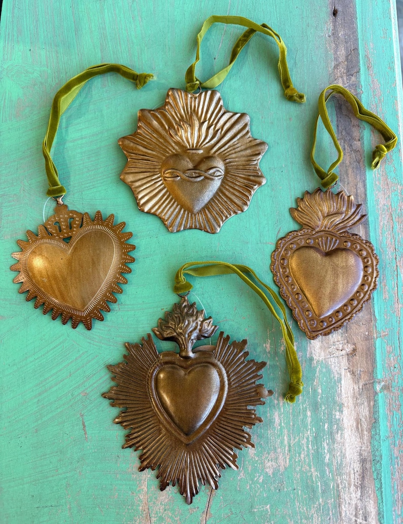 K&ouml;nnte beinhalten: Vier Metallherz-Ornamente im antiken Stil mit Gold-Finish, jedes mit einem einzigartigen Design und mit gr&uuml;nem Samtband befestigt. Die Ornamente werden auf einer verwitterten t&uuml;rkisfarbenen Holzoberfl&auml;che pr&auml;sentiert.