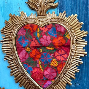 以下が含まれることがあります： 金色のフレームとカラフルな花のテキスタイルセンターを備えた、ハート型の壁掛け。 フレームはサンバーストのデザインと装飾的な上部要素を備えています。 テキスタイルには、赤、ピンク、オレンジ、青の花が描かれています。