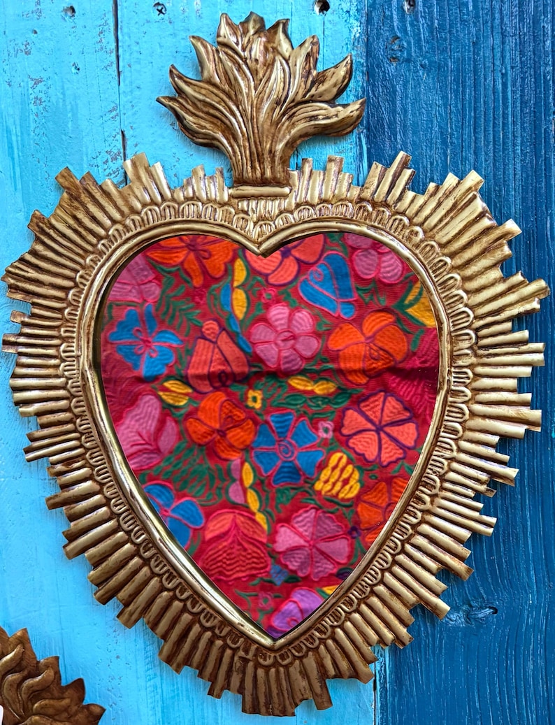 以下が含まれることがあります： 金色のフレームとカラフルな花のテキスタイルセンターを備えた、ハート型の壁掛け。 フレームはサンバーストのデザインと装飾的な上部要素を備えています。 テキスタイルには、赤、ピンク、オレンジ、青の花が描かれています。