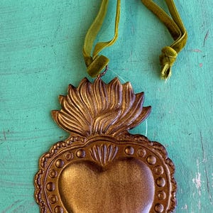K&ouml;nnte beinhalten: Ein bronzefarbenes herzf&ouml;rmiges Ornament mit dekorativem Rand und floralem Aufsatz. Das Ornament ist ca. 10 cm x 6,3 cm gro&szlig; und hat ein gr&uuml;nes Samtband zum Aufh&auml;ngen. Der Text "Small Dots" wird auf orangefarbenem Hintergrund angezeigt.