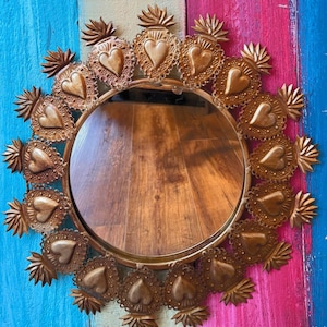 May include: A circular mirror with a gold-toned metal frame. The frame is adorned with a repeating pattern of heart-shaped motifs and flame-like details. The mirror is set against a backdrop of vertical stripes in blue, pink, and green.