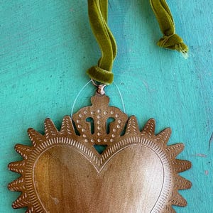 K&ouml;nnte beinhalten: Ein kleiner, goldfarbener Metallherzanh&auml;nger, 7,6 cm x 8,9 cm, mit Kronendetail und Sonnenstrahlenrand. Das Herz wird von einem gr&uuml;nen Samtband gehalten. Der Text "Small Crowned Heart (3" x 3.5")" befindet sich in einer orangefarbenen Box.