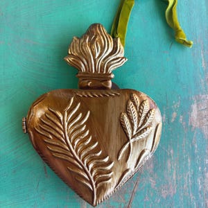 May include: A heart-shaped, gold-colored metal ornament with embossed leaf designs and a decorative top. A green velvet ribbon is attached for hanging. The ornament is displayed on a distressed turquoise surface.