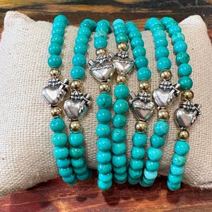 Peut inclure: Six bracelets de perles turquoise avec des breloques en forme de cœur argentées et des accents dorés sont présentés sur un coussin beige. Les bracelets sont faits de petites perles rondes et sont probablement extensibles. Les breloques en forme de cœur ont un design détaillé.