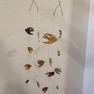 Puede incluir: Móvil decorativo con siluetas de pájaros dorados suspendidas de un marco metálico curvo. Los pájaros están suspendidos por finos hilos, creando un diseño delicado y aéreo. Ideal para la decoración interior.