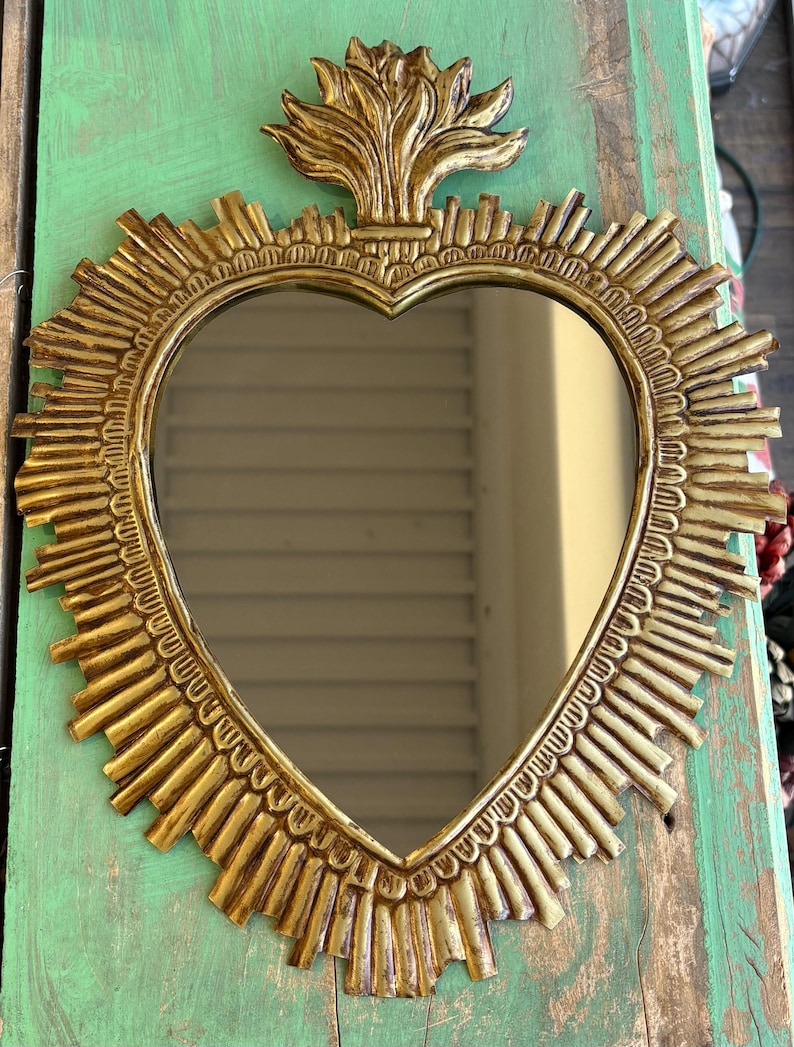以下が含まれることがあります： 縁にサンバーストデザインが施された、ハート型の金色の鏡。鏡には装飾的な上部要素があります。鏡は、風化した緑色の木製の表面に表示されています。