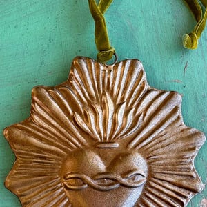 K&ouml;nnte beinhalten: Ein metallisch goldfarbener, runder Heiliges Herz-Ornament, ca. 10 cm x 10 cm, mit einem Sonnenstrahl-Design. Das Herz zeigt Flammen und eine Dornenkrone. Es h&auml;ngt an einem gr&uuml;nen Samtband.
