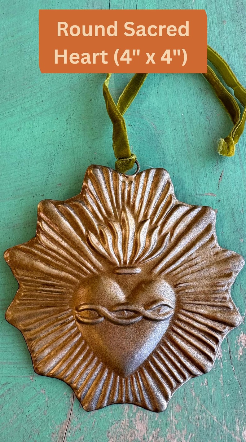 K&ouml;nnte beinhalten: Ein metallisch goldfarbener, runder Heiliges Herz-Ornament, ca. 10 cm x 10 cm, mit einem Sonnenstrahl-Design. Das Herz zeigt Flammen und eine Dornenkrone. Es h&auml;ngt an einem gr&uuml;nen Samtband.
