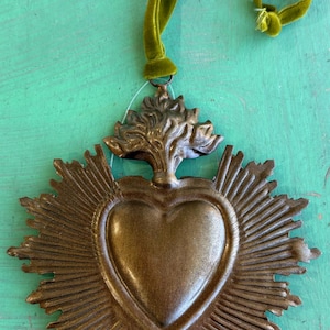 K&ouml;nnte beinhalten: Bronze-farbener Metall-Wandbehang in Herzform mit Sonnenstrahlen-Design, 12,7 cm x 10,2 cm. Das Herz ist mit einem baumartigen Design gekr&ouml;nt und hat ein gr&uuml;nes Samtband zum Aufh&auml;ngen. Der Text "Small Rays" befindet sich in einem orangefarbenen Rechteck.