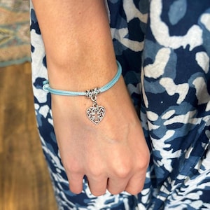 May include: A pale blue cord bracelet featuring a silver heart-shaped charm. The charm has a cross design. The bracelet is secured with a knot.