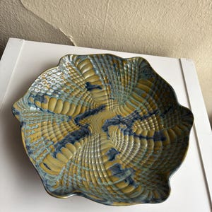 May include: A decorative ceramic bowl with a unique, textured design. The bowl features a wavy rim and a pattern of intersecting lines in shades of yellow, blue, and green. The bowl's surface has a tactile, ridged texture.
