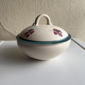 Könnte beinhalten: Eine cremefarbene Keramikschüssel mit Deckel und Griff. Der Deckel hat einen türkisfarbenen Rand und rote Blumendesigns. Die Schüssel ist rund und steht auf einer weißen Oberfläche. Der Griff ist gewölbt.