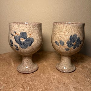 Pode incluir: Dois cálices de cerâmica salpicada com um desenho floral. Os cálices têm uma cor de base bege com detalhes florais azuis. A haste e a base são de cor bege lisa. Os cálices são exibidos em uma superfície marrom.