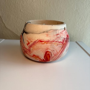 May include: A small, round ceramic pot with a wide opening. The exterior has a marbled design in red, orange, and white. The rim is a natural, unglazed colour. The pot is likely used for decorative purposes, perhaps for holding small items.