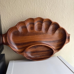 May include: A wooden serving tray with a scalloped edge and two sections. The tray is made of a dark brown wood with visible grain patterns. It has a large, oval-shaped section and a smaller, elongated section. The tray has two handles.