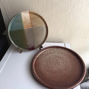 Puede incluir: Dos platos de cerámica hechos a mano. El plato superior está dividido en cuatro secciones de verde, beige y marrón. El plato inferior es de color marrón liso con una textura moteada. Ambos platos son redondos y tienen una estética rústica.