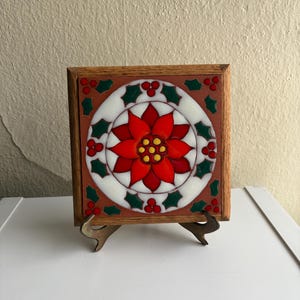 May include: Square decorative tile with a poinsettia design. The tile features a red flower with yellow center, surrounded by green holly leaves and red berries, set against a white and brown background. The tile is framed in wood and displayed on a metal stand.
