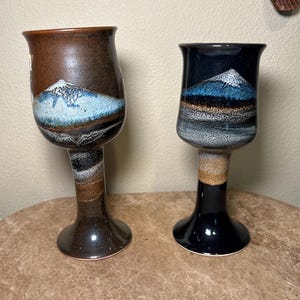 May include: Two ceramic goblets with mountain landscape designs. One goblet is brown with a blue and white mountain scene, the other is black with a similar design. Both have a flared rim and a pedestal base.
