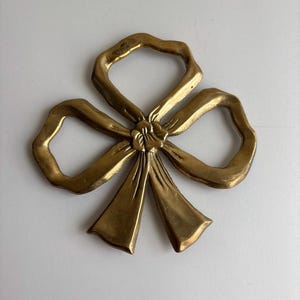 May include: A gold-coloured metal bow-shaped decorative object. The bow has three loops and a central knot with flowing ribbon ends. The metal has a slightly textured surface and a warm, antique finish. The object is likely a decorative element.