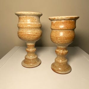 Puede incluir: Dos copas de cerámica hechas a mano con un aspecto rústico y texturizado. Las copas son de color beige cálido con rayas horizontales marrones y un borde acampanado. Tienen una base de pedestal y un cuerpo bulboso.