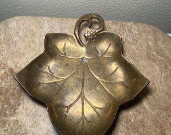 May include: A vintage brass leaf-shaped dish with a detailed vein pattern. The dish has a small handle with a cluster of grapes. The brass has a warm, aged patina, adding to its antique charm. This decorative piece is ideal for holding small items.