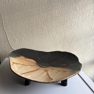 May include: A handmade ceramic serving dish with a unique, organic shape. The dish features a dark brown center that fades to tan and green edges. It is supported by three small, black legs, adding a touch of elegance to this decorative piece.