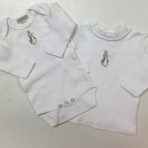 Könnte beinhalten: Zwei weiße Langarmshirts mit Peter Rabbit Stickerei. Ein Shirt ist ein Body mit drei Druckknöpfen am unteren Rand. Das andere Shirt hat einen Rundhalsausschnitt.